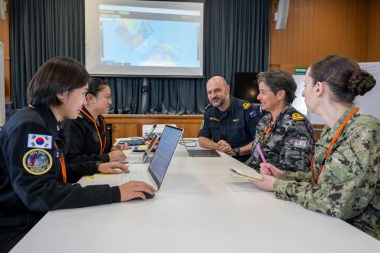 Four women and one man in naval uniform seated around a table with laptops.