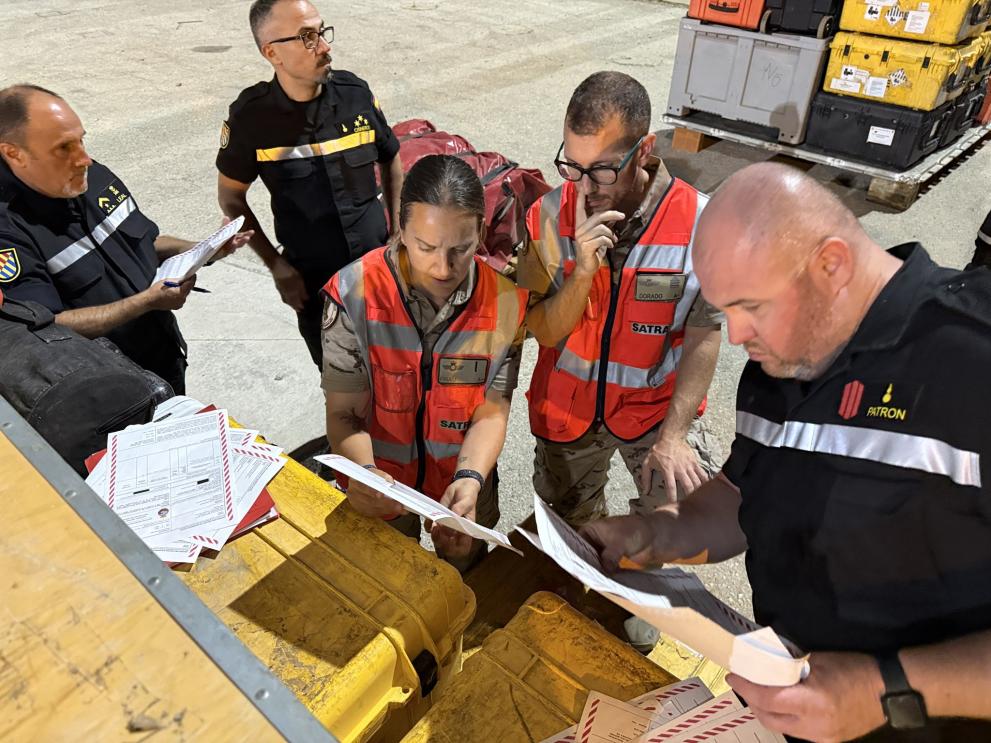 A group of emergency response workers in uniforms and high-visibility vests stand around large yellow equipment cases at night, reviewing and sorting paperwork beside stacks of packed crates, suggesting coordination of a humanitarian or disaster relief operation.