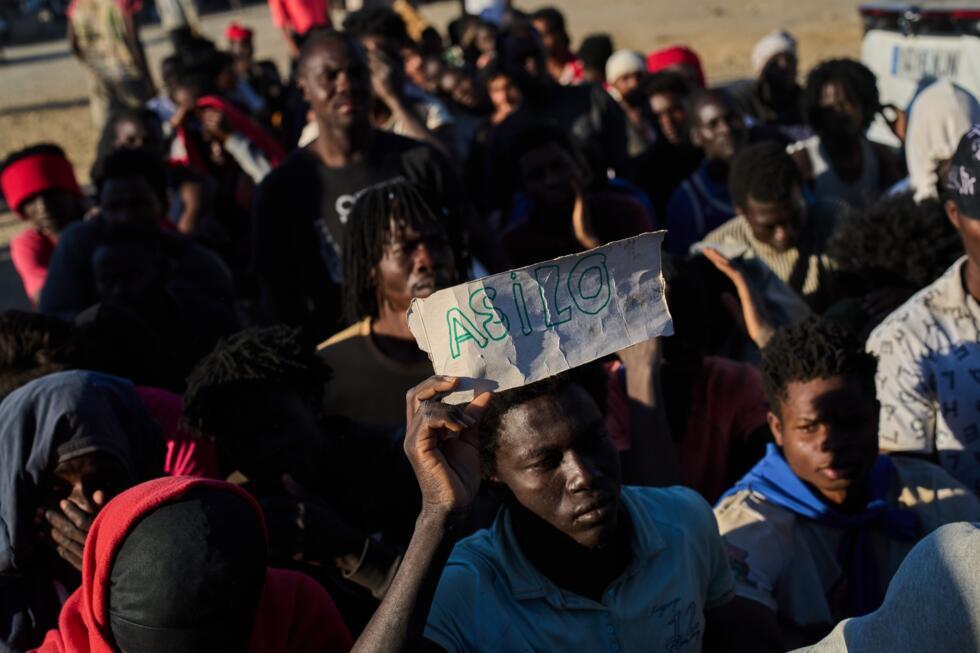 A migrant who crossed from Morocco into Spain holds a sign reading "asylum" in Spanish as he lines up for food distribution on a beach in the Spanish enclave of Ceuta on Monday, August 3, 2026
